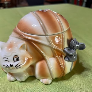 Puede incluir: Un recipiente de cerámica con forma de gato con tapa, con una cara guiñando un ojo y una figura de ratón gris agarrada al costado. El recipiente es principalmente naranja y blanco, con un acabado brillante. Un artículo decorativo.
