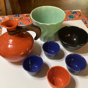 May include: A set of vintage ceramic kitchenware in various colors, including a red pitcher with a wooden handle, a green planter, a black bowl, and four small bowls in blue and orange.