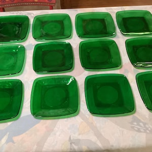 Può includere: Una collezione di dodici piatti quadrati in vetro verde smeraldo. Ogni piatto ha una forma quadrata leggermente arrotondata con un'indentazione circolare centrale e un motivo decorativo. I piatti sono disposti su una superficie bianca.