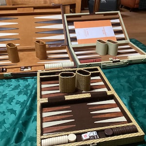 Puede incluir: Tres juegos de backgammon abiertos se exhiben sobre un mantel verde. Los juegos presentan superficies de juego marrones, beige y blancas, con dados, fichas y cubiletes. Los juegos son en tonos marrones y beige, algunos con detalles en verde claro.