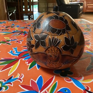 May include: A handcrafted gourd with a brown finish, featuring intricate carvings of flowers and an owl. The gourd is set on a vibrant orange tablecloth with a colorful floral pattern, creating a striking contrast.