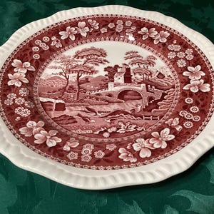 May include: A round, white and red decorative plate with a scalloped edge. The plate features a detailed landscape scene in the centre, surrounded by floral and geometric patterns. The plate measures approximately 25 cm in diameter.