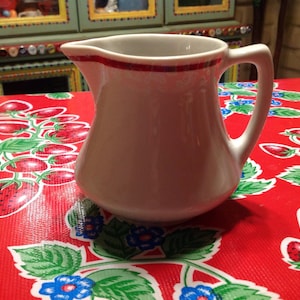 May include: A white ceramic creamer with a red rim and a handle. The creamer is sitting on a red tablecloth with a strawberry pattern.