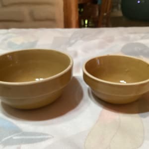 May include: Two ceramic bowls, one larger than the other, are shown on a white tablecloth. Both bowls are a light brown colour.