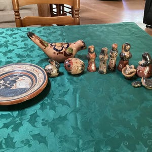 May include: A collection of hand-painted ceramic figurines and a decorative plate. The figurines depict various figures with detailed patterns in shades of red, brown, and black. The plate features a bird design with blue and brown accents.