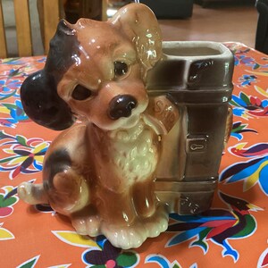 May include: A ceramic dog planter with a brown and white color scheme. The dog is sitting next to a brown mailbox-shaped planter. The word "Said" is on a sign attached to the mailbox. The planter is on a colorful floral tablecloth.
