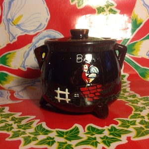 Puede incluir: Un tarro de cerámica marrón oscuro con tapa, dos asas y tres patas. El tarro presenta una ilustración de gallo con la palabra "B" y un diseño de chimenea. El tarro está sobre un mantel rojo con un estampado floral.