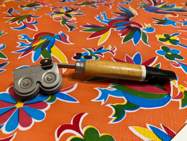 May include: A vintage can opener with a wooden handle and a silver metal cutting mechanism. The opener has two wheels with serrated edges for opening cans.