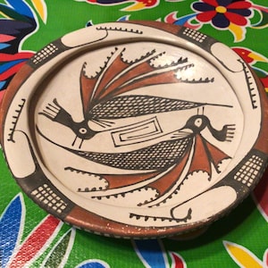 Puede incluir: Un plato de cerámica blanco con un diseño rojo, negro y blanco de dos pájaros estilizados con las alas extendidas. El plato tiene un borde marrón.