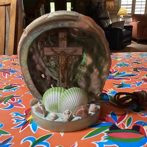 May include: A decorative lamp made from a large seashell with a cross inside. The shell is iridescent and has a green and white interior. The base of the lamp is decorated with small seashells.