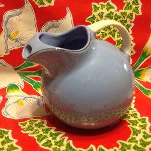 May include: A light blue ceramic creamer with a white rim and handle. The creamer is sitting on a red tablecloth with white flowers and green leaves.