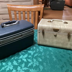 Könnte beinhalten: Zwei Vintage-Reisekoffer. Einer ist marineblau mit weißem Rand und schwarzem Griff. Der andere ist hellbeige mit messingfarbener Hardware und einem Griff. Beide Koffer sind rechteckig und stehen auf einer grünen Tischdecke.