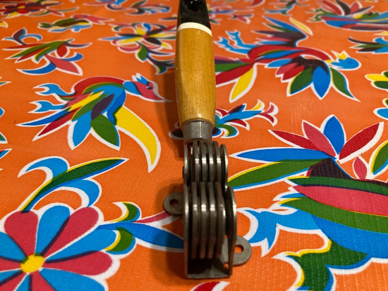 May include: A vintage knife sharpener with a wooden handle and a metal sharpening mechanism. The handle is black and white with a wood grain pattern. The sharpening mechanism has multiple metal blades.