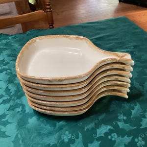 May include: A stack of seven square ceramic dishes with handles. Each dish features a smooth white interior and a textured, light tan, marbled rim. The dishes are uniform in size and shape, designed with a single handle extending from one side.