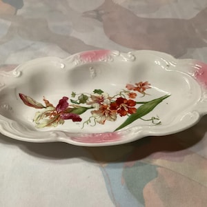 May include: An oval, white ceramic serving dish with a scalloped edge and pink accents. The dish features a floral design with red, pink, and green flowers. The dish is approximately 30 cm long.
