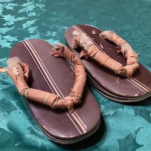 May include: A pair of traditional Japanese geta sandals. The wooden platforms are dark brown with white and gold stripes. The straps are made of a worn, tan fabric. The sandals are elevated and show signs of age.