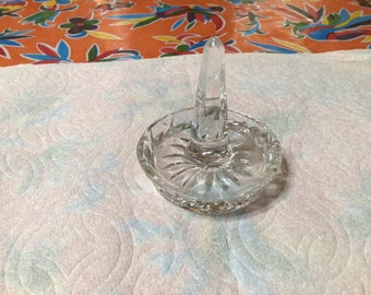 Op de afbeelding: Een glazen ringhouder van helder glas met een verhoogde middelste paal en een decoratieve, geslepen glazen basis.