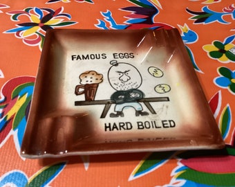 Vintage MCM square ceramic comical ashtray with saying, “Famous eggs, hard boiled” and a mean looking egg.