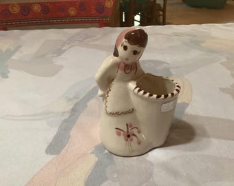 Puede incluir: Figura de cerámica vintage de una niña sosteniendo una cesta. La figura está pintada en tonos crema, rosa y marrón. La niña lleva un gorro y un delantal, y la cesta tiene un borde decorativo. Un diseño floral está pintado en la parte delantera.