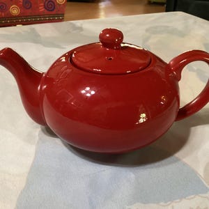 May include: A vibrant red ceramic teapot with a lid and a curved handle. The teapot has a rounded body, a spout, and a small knob on the lid. This kitchenware item is designed for brewing tea.