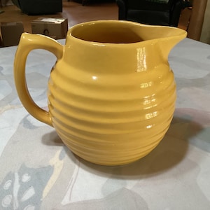 May include: A yellow ceramic jug with a handle and a spout. The jug has a ribbed design around the body. The jug is on a table.