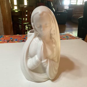 Op de afbeelding: Een witte keramieken beeldje van een vrouw in een gedrapeerd gewaad, met haar hoofd gebogen en handen gevouwen in gebed. Het beeldje heeft een gladde, matte afwerking en is ongeveer 20 cm hoog.