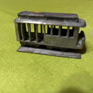 Puede incluir: Un pequeño juguete de tranvía de metal antiguo. El tranvía de color plateado tiene un diseño detallado con barras verticales para las ventanas y un techo. El juguete se asienta sobre una base rectangular y tiene ruedas.