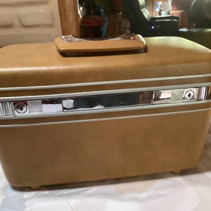 Könnte beinhalten: Ein vintage brauner Hartschalen-Reisekoffer mit einem silbernen Metallgriff und zwei Schlössern. Der Koffer hat ein hellbraunes Innenfutter und einen weißen Rand an den Kanten.
