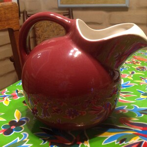 May include: A maroon ceramic pitcher with a white rim and a handle. The pitcher has a round body and a slightly curved spout. It is sitting on a green tablecloth with a colorful floral pattern.