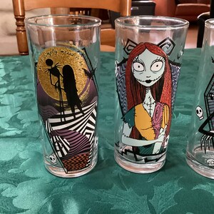 Puede incluir: Tres vasos de cristal transparente con personajes de Pesadilla antes de Navidad. Un vaso muestra a Jack y Sally ante una luna de purpurina dorada con el texto 'We're simply meant to be'. Otro vaso muestra a Sally y el tercero a Jack.