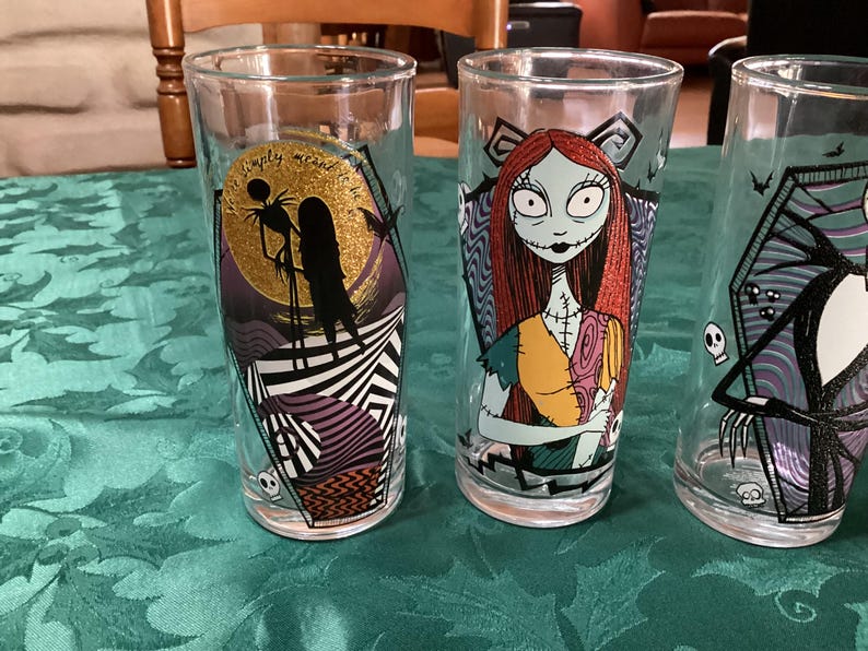 Puede incluir: Tres vasos de cristal transparente con personajes de Pesadilla antes de Navidad. Un vaso muestra a Jack y Sally ante una luna de purpurina dorada con el texto 'We're simply meant to be'. Otro vaso muestra a Sally y el tercero a Jack.