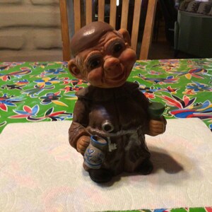 Peut inclure: Une figurine en céramique brune d'un moine tenant une tasse verte dans une main et une tasse brune dans l'autre. Le moine porte une robe brune et une ceinture en corde blanche. La figurine est sur une surface blanche.