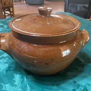May include: A brown ceramic casserole dish with a lid. The dish has two handles and a knob on the lid. The dish is sitting on a teal tablecloth. The casserole dish is a kitchenware item.