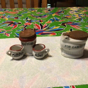 Puede incluir: Un juego de cuatro tarros de cerámica vintage en forma de inodoro. El tarro más grande está etiquetado como "Solo para damas". Los otros tres tarros están etiquetados como "Cigarrillos", "Cerillas" y "Ceniza".