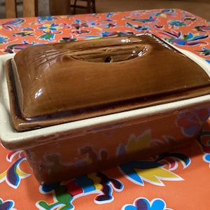 Può includere: Una pirofila in ceramica marrone con coperchio. La pirofila ha un bordo bianco e un motivo floreale sul fondo. Il coperchio ha un piccolo foro al centro.