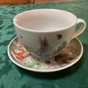 May include: A white ceramic teacup and saucer set featuring a colorful illustration of a rabbit wearing a blue jacket in a garden setting. The saucer displays detailed botanical imagery and additional small rabbit figures in red clothing.