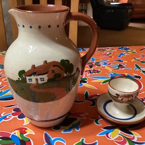 Może przedstawiać: Biały ceramiczny dzbanek z brązowym uchwytem i malowanym motywem domu z czerwonym dachem i zielonymi drzewami. Dzbanek stoi na kolorowym obrusie. Jest też mały biały ceramiczny kubek na jajka z malowanym motywem domu z czerwonym dachem i zielonymi drzewami. Kubek na jajka stoi na białym ceramicznym spodku z niebieskim i brązowym rantem.