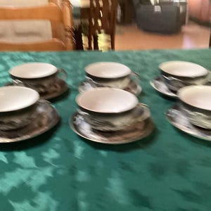 May include: A set of six ceramic teacups and matching saucers featuring a dark, metallic-toned exterior with a white interior. Each cup has a curved handle and sits on a circular saucer, arranged in two rows on a green surface.