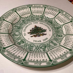 May include: A white ceramic plate with a green border and calendar months encircling the edge. The center features a Christmas tree design with the text "To Celebrate the Millennium Year 2000".