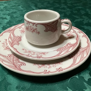 Könnte beinhalten: Ein weißes Keramik-Teeservice mit rotem Blumendesign. Das Set besteht aus einer Teetasse, einem kleinen Teller und einem größeren Teller. Die Teller und die Tasse haben einen gewellten Rand.