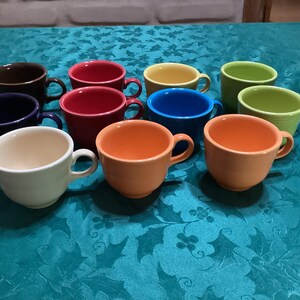 Op de afbeelding: Een verzameling kleurrijke keramische koffiekopjes in verschillende tinten, waaronder bruin, rood, blauw, geel, groen, oranje en wit. Elk kopje heeft een afgeronde vorm met een oor. De kopjes staan op een turquoise tafelkleed.