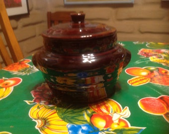 Puede incluir: Una olla de cerámica marrón con tapa, que presenta un diseño floral en tonos amarillos, verdes y azules. La olla está sentada sobre un mantel verde con un patrón de frutas.