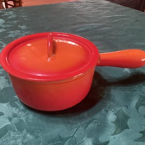 May include: A small, orange enameled cast iron pot with a lid and a handle. The pot is round with a matching lid and a single, curved handle. The pot sits on a green tablecloth with a holly leaf pattern.