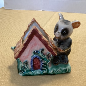 Puede incluir: Una figurita de cerámica de un animal pequeño, posiblemente un tejón, sosteniendo una casa rosa y marrón con un techo verde y una puerta roja. La figurita está pintada con detalles en verde, azul y marrón.