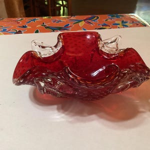May include: A red glass bowl with a textured surface and a ruffled edge. The bowl is made of clear glass with red glass bubbles throughout.