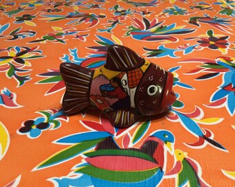 Pode incluir: Uma figurinha de peixe de cerâmica colorida com um corpo castanho, detalhes amarelos, vermelhos e azuis, e um olho branco. O peixe está sentado sobre uma toalha de mesa laranja brilhante com um padrão floral.