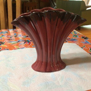 Puede incluir: Un jarrón de cerámica granate con un diseño acanalado. El jarrón tiene una parte superior ancha y con volantes y una base acampanada. La parte superior del jarrón es de un tono granate más oscuro. El jarrón está sobre un mantel naranja y blanco estampado.