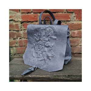 Op de afbeelding: Een grijze leren rugzak met een geborduurd bloemenpatroon aan de voorkant. De rugzak heeft een klep sluiting en een gespsluiting.