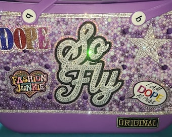 May include: A lilac tote bag with a sparkly design. The bag has rhinestone lettering spelling "DOPE" and "So Fly", a star, and the words "Fashion Junkie". The bottom of the bag reads "ORIGINAL".