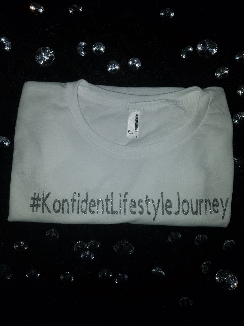 Puede incluir: Camiseta blanca con el texto "#KonfidentLifestyleJourney" impreso en purpurina plateada.